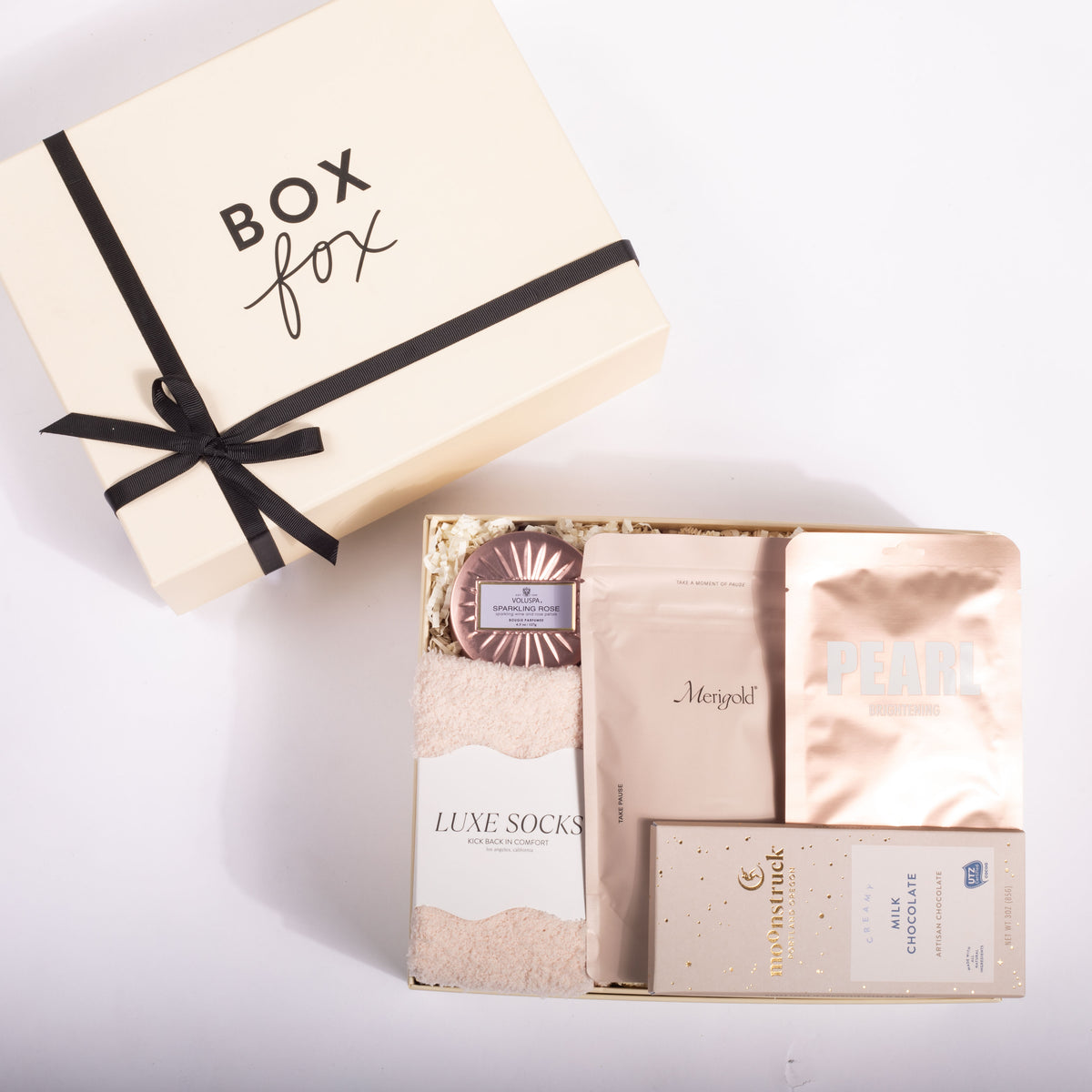 A luxury gift set from Box Fox, featuring a cream-colored box with a black ribbon next to an open box containing skincare and comfort items including Pearl Face Mask, Luxe Socks, and a chocolate bar, on a white background.