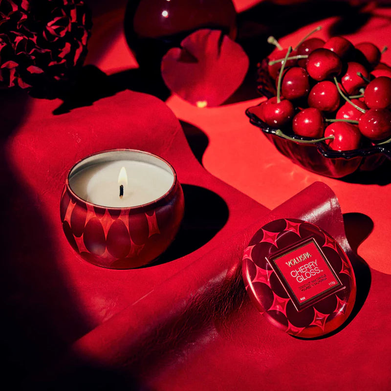 Candle in a red holder with a small container labeled 'Waxwork Cherry Gloss' on a red surface.