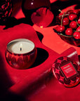 Candle in a red holder with a small container labeled 'Waxwork Cherry Gloss' on a red surface.