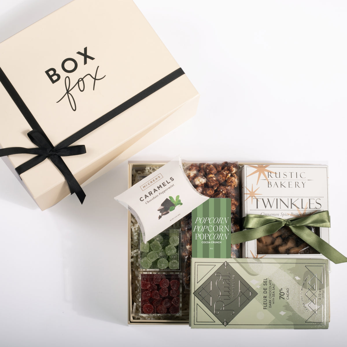 A luxury gourmet gift box set from Box Fox, featuring a cream-colored gift box with a black ribbon and a tray of assorted gourmet snacks including caramel-coated chocolates, popcorn, dried cranberries, and Twinkles, arranged on a clean white background.