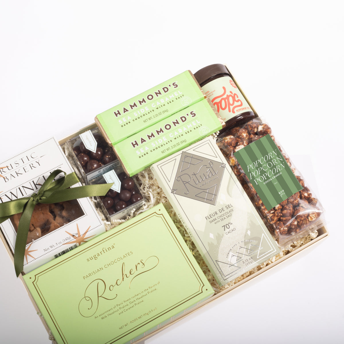 A curated gourmet gift box filled with assorted packaged snacks and chocolates, including Hammond's dark chocolate bars, a jar of Poppy Seed Butter, and a bag of Roach's Persian Chocolate, arranged in a light wood box with a green ribbon on a white background.
