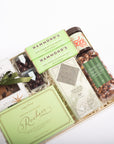 A curated gourmet gift box filled with assorted packaged snacks and chocolates, including Hammond's dark chocolate bars, a jar of Poppy Seed Butter, and a bag of Roach's Persian Chocolate, arranged in a light wood box with a green ribbon on a white background.
