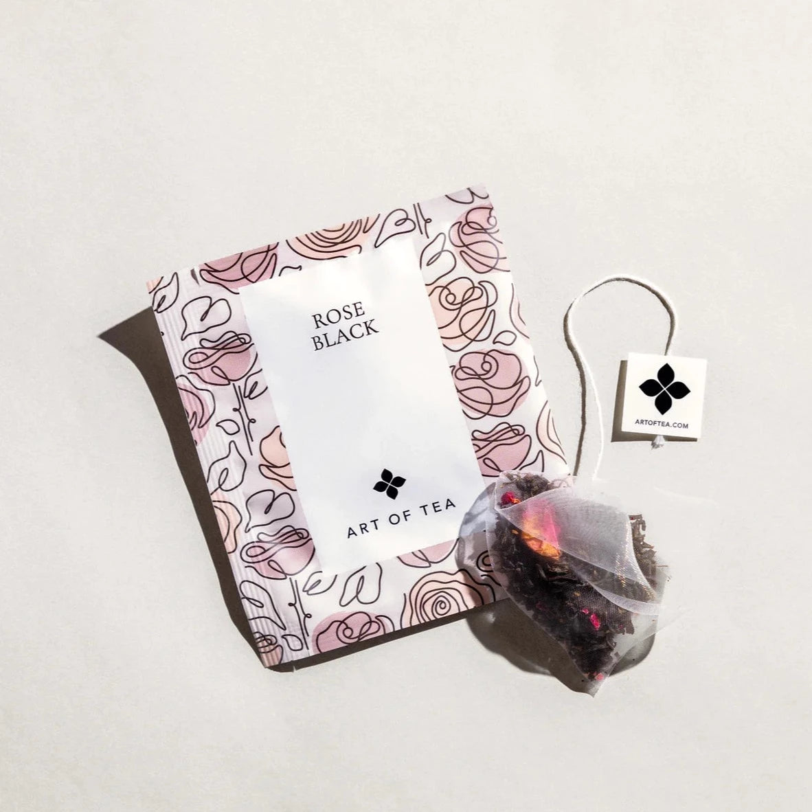 Rose Black tea sachet. Sachet has doodles of roses all over it and sachet is a very pale pink. Each doodled rose has a various shade of pink. Tea bag is outside the sachet
