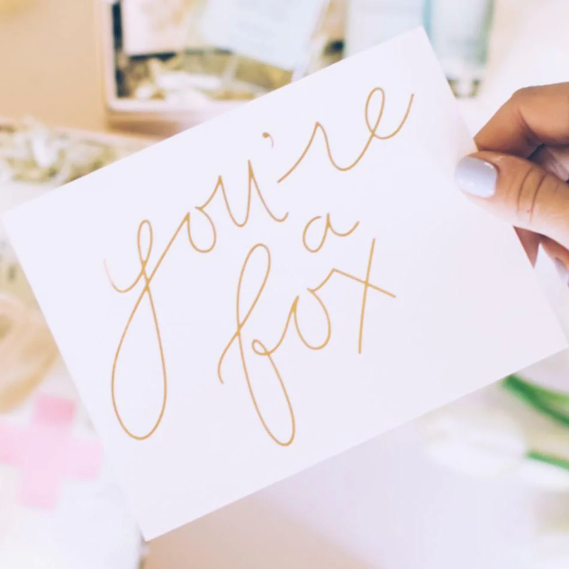 White card that reads "you're a fox" in gold foil script. Thumb with blue polish holds the card at an angle.