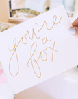 White card that reads "you're a fox" in gold foil script. Thumb with blue polish holds the card at an angle.