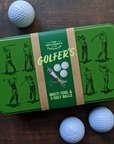 GREEN TIN CONTAINER LABELED "GOLFER'S MULTI-TOOL & 3 GOLF BALLS" IN WHITE ON A BROWN WOOD TABLE NEXT TO THREE WHITE GOLF BALLS