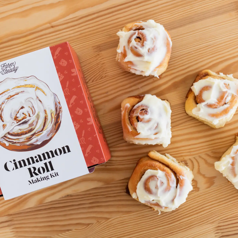 Cinnamon Roll making kit that has red and white packaging. There are cinnamon rolls next to the packaging and they all are on a wooden table.