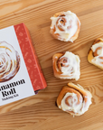 Cinnamon Roll making kit that has red and white packaging. There are cinnamon rolls next to the packaging and they all are on a wooden table.