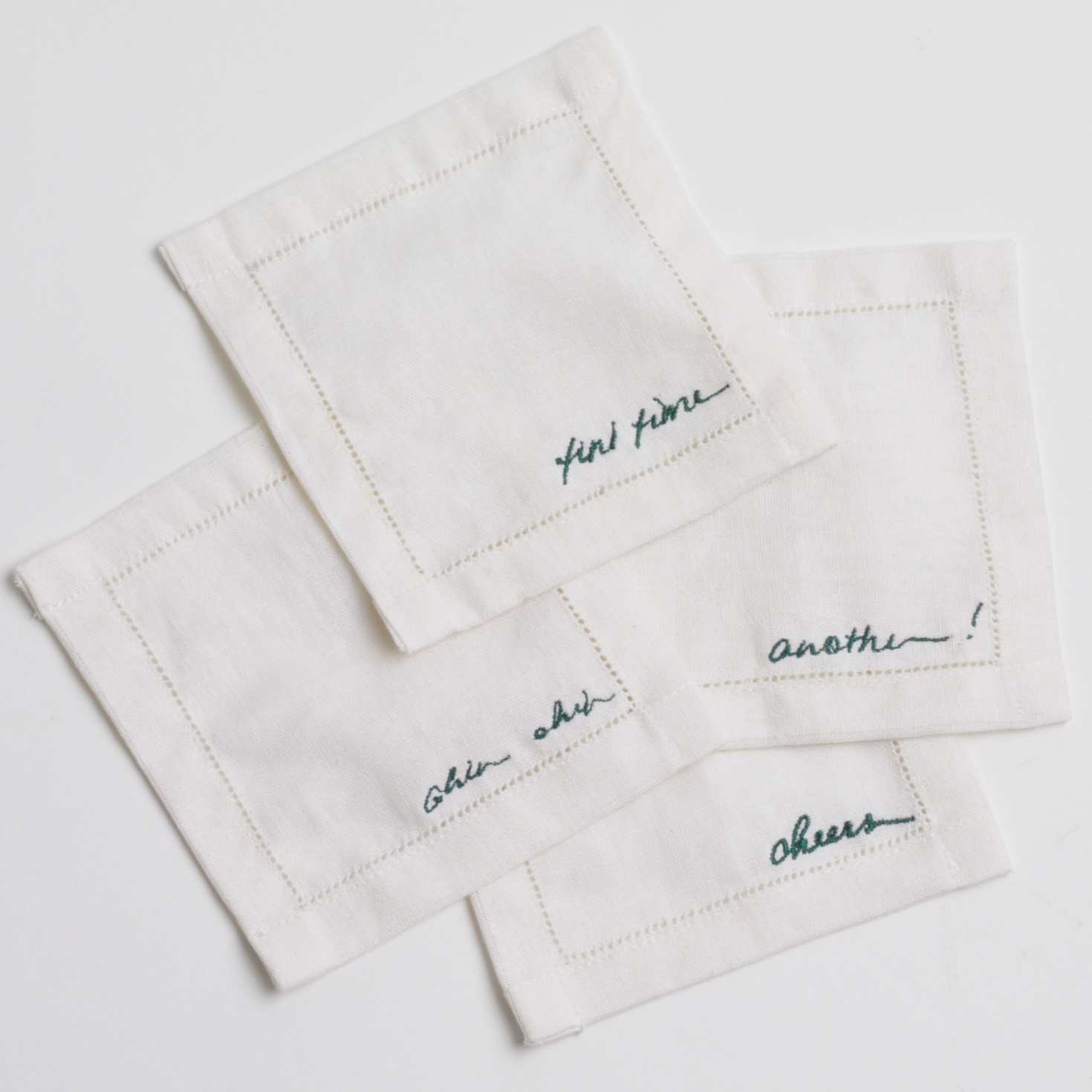 A set of four cocktail napkins with embrodiered script on them behind white background 