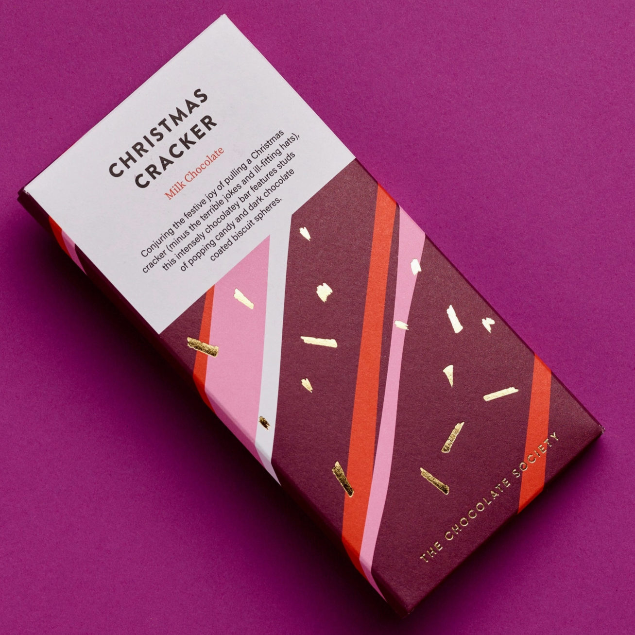 A stylishly designed Christmas Cracker chocolate bar by The Chocolate Society, placed on a magenta background. The packaging features a white section with product details and a festive design with pink, red, and gold diagonal stripes against a maroon base, accented by gold foil confetti. The packaging highlights that it contains milk chocolate.