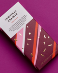 A stylishly designed Christmas Cracker chocolate bar by The Chocolate Society, placed on a magenta background. The packaging features a white section with product details and a festive design with pink, red, and gold diagonal stripes against a maroon base, accented by gold foil confetti. The packaging highlights that it contains milk chocolate.