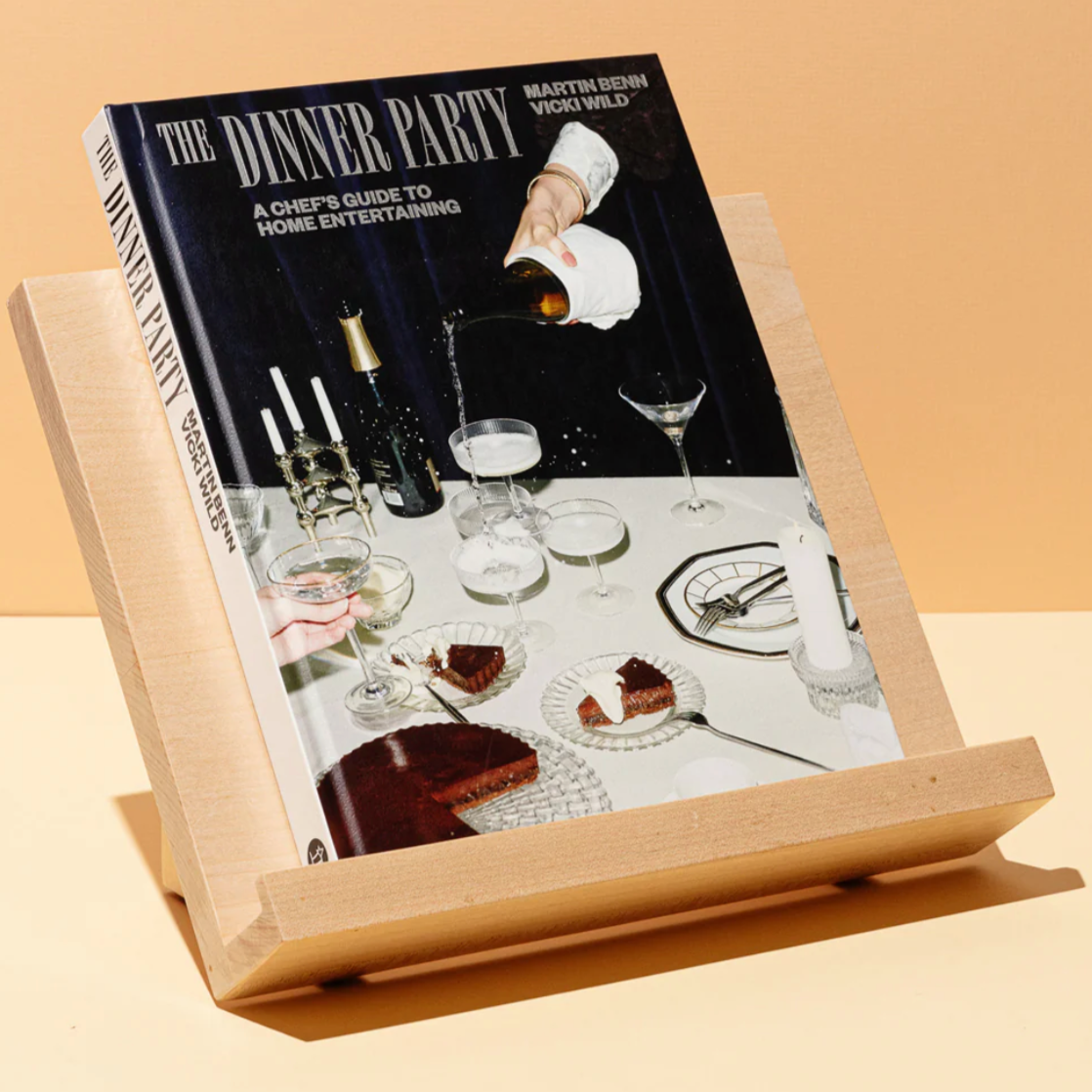 On an orange and white background, the Dinner Party Book sits propped up. The cover is shown- it is a dinner table with coupe glasses and cake.