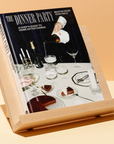 On an orange and white background, the Dinner Party Book sits propped up. The cover is shown- it is a dinner table with coupe glasses and cake.