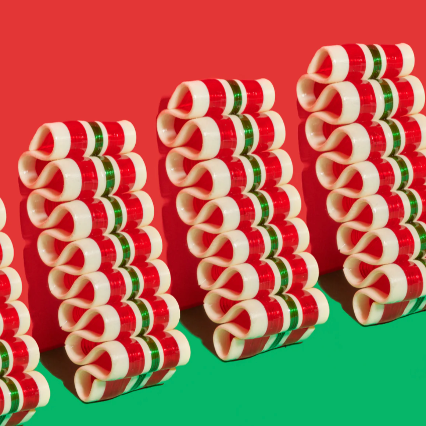 The image shows rows of neatly stacked cinnamon candy bundles in a spiral shape, alternating in red, white, and green colors. The background is split diagonally, with a festive red upper half and a green lower half, complementing the holiday-themed colors of the candy. 