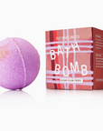 This image shows a purple bath bomb next to its packaging. The bath bomb has a spherical shape with a slight glittery texture on one side, giving it a shimmery look. The packaging is a rectangular box with a striped design in shades of red, pink, and white. The text on the box reads "Old Whaling Co. Bath Bomb" with the scent labeled as "Sugar Plum Ferry." There are also small sections of text on the side of the box providing instructions and information about the product.






