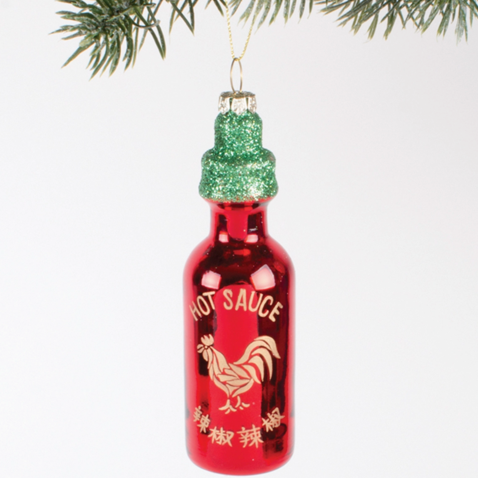 red hot sauce bottle christmas ornament with green glittery cap hanging behind white background