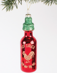 red hot sauce bottle christmas ornament with green glittery cap hanging behind white background