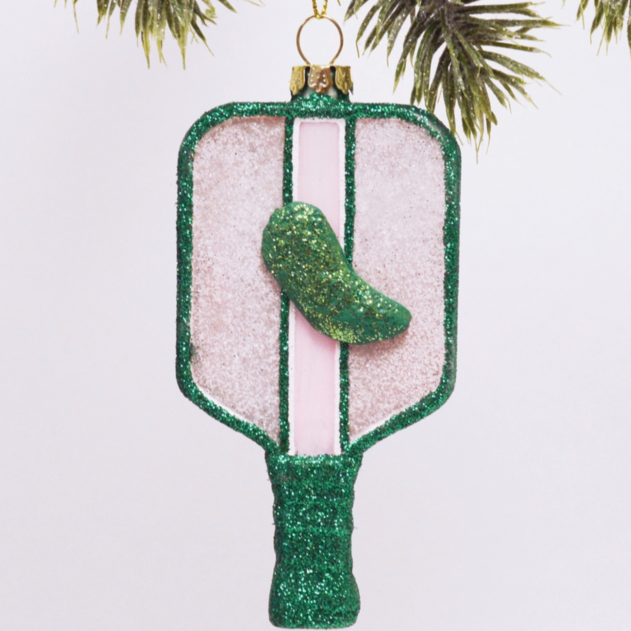 this image shows a pickleball racket shaped ornament, it is glittery with a green glittery pickle in the middle, green light pinks and whites are the main colors 