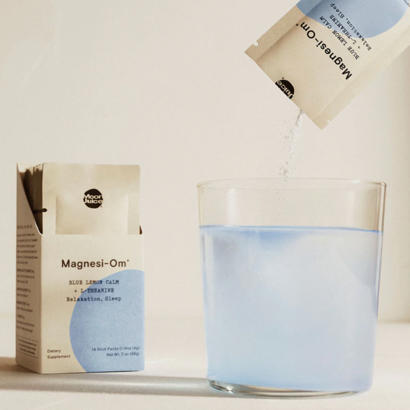 The image shows a product named "Magnesi-Om" by Moon Juice. On the left, a box of individual sachets is displayed, labeled "Magnesi-Om" with a subtitle that reads "Blue Lemon Calm + L-Theanine" and mentions benefits such as "Relaxation, Sleep." One sachet is open and appears to be pouring powder into a clear glass of water on the right. The water is turning light blue, likely from the powder dissolving. The scene is minimalist with neutral tones, emphasizing the product.


