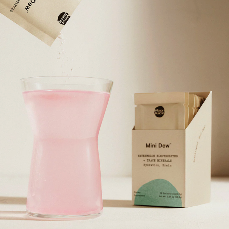 The image shows a product called "Mini Dew" by Moon Juice. It features a box with individual sachets next to a glass filled with a light pink liquid, created by pouring the contents of a sachet into water. The sachet is labeled "Watermelon Electrolytes + Trace Minerals," indicating the product's purpose of providing hydration and brain support. The powder is being poured into the glass, and the pink color of the liquid suggests a watermelon flavor. 