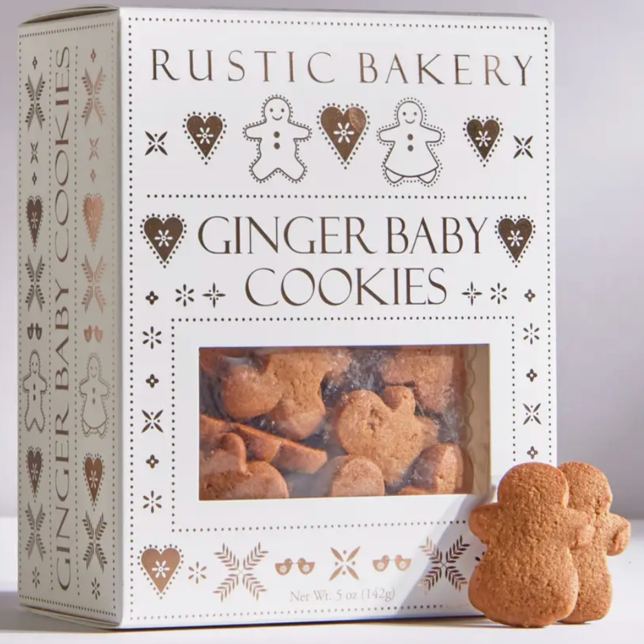 White and gold box of ginger baby cookies by rustic bakery with two cookies outside of the box to show the shape. cookies are shaped like mini ginger bread men. behind grey background.