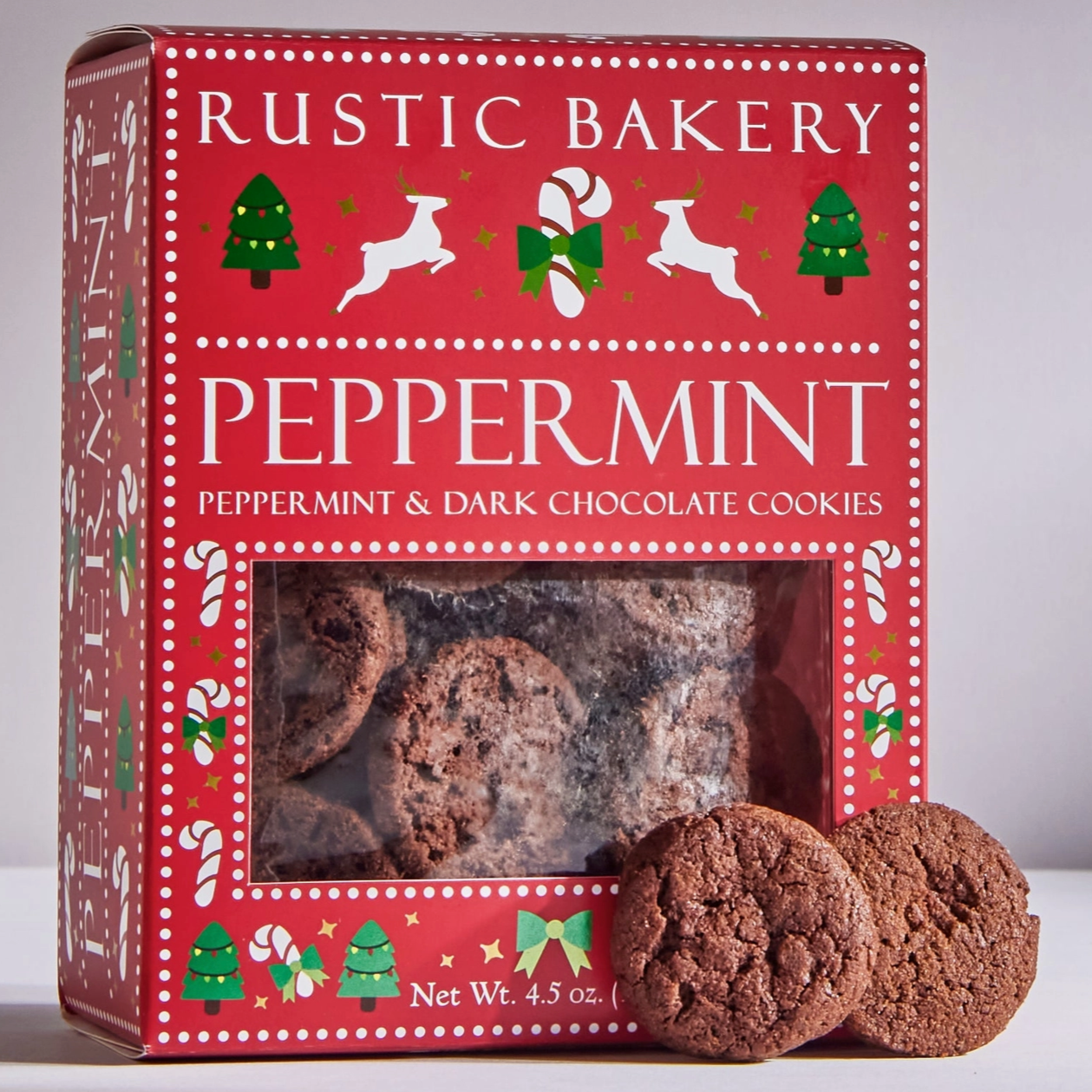 Rustic Bakery red and green box of peppermint & dark chocolate cookies. reindeer, candy canes and christmas trees are decorated on the box and 2 ginger dark chocolate cookies are outside to the right of the box to display the shape and look of the cookie behind white/grey background