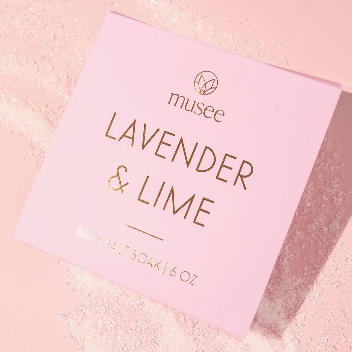 light lavendar packaging of musee 6 oz lavendar & lime bath salt soak in front of light pink background with visible salt. 