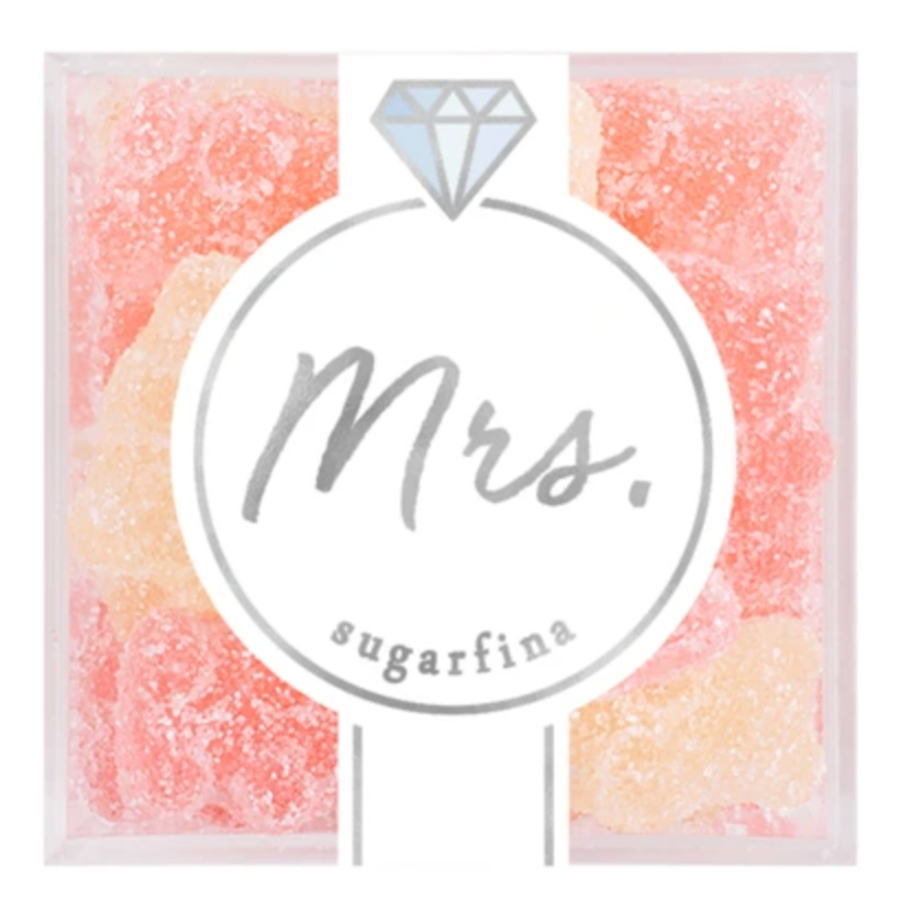 The image shows a clear box of gummy candies by Sugarfina, labeled "Mrs." The front of the packaging has a minimalist design featuring a stylized diamond icon above the word "Mrs." The gummies inside are dusted with sugar and come in shades of light pink and peach, suggesting a sweet, delicate flavor. The packaging design is elegant and likely intended for celebratory or bridal-themed occasions.