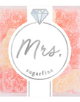 The image shows a clear box of gummy candies by Sugarfina, labeled "Mrs." The front of the packaging has a minimalist design featuring a stylized diamond icon above the word "Mrs." The gummies inside are dusted with sugar and come in shades of light pink and peach, suggesting a sweet, delicate flavor. The packaging design is elegant and likely intended for celebratory or bridal-themed occasions.