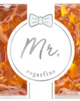 The image shows a package of gummy candies by Sugarfina. The packaging design has a label in the center with the text "Mr." written in a gray, cursive font. Above the text, there's a small, simple bow-tie graphic. Surrounding the label, the gummies are visible through the clear packaging. The gummies are amber-colored and shaped like bears.
