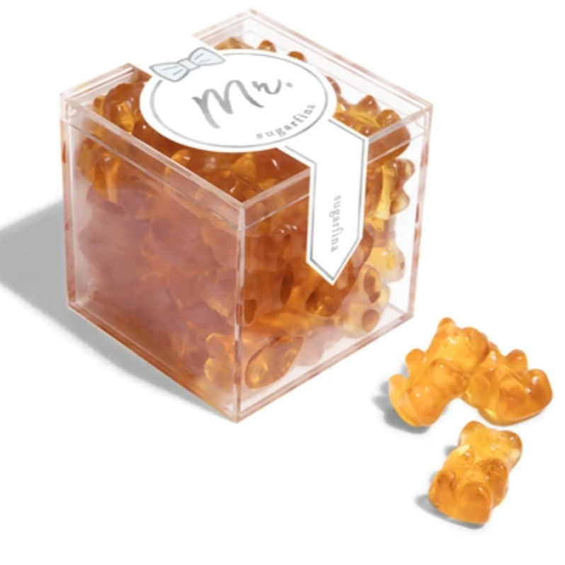 The image shows a package of gummy candies by Sugarfina. The packaging design has a label in the center with the text "Mr." written in a gray, cursive font. Above the text, there's a small, simple bow-tie graphic. Surrounding the label, the gummies are visible through the clear packaging. The gummies are amber-colored and shaped like bears. There are three bears outside of the box pictured to the right of the box. 





