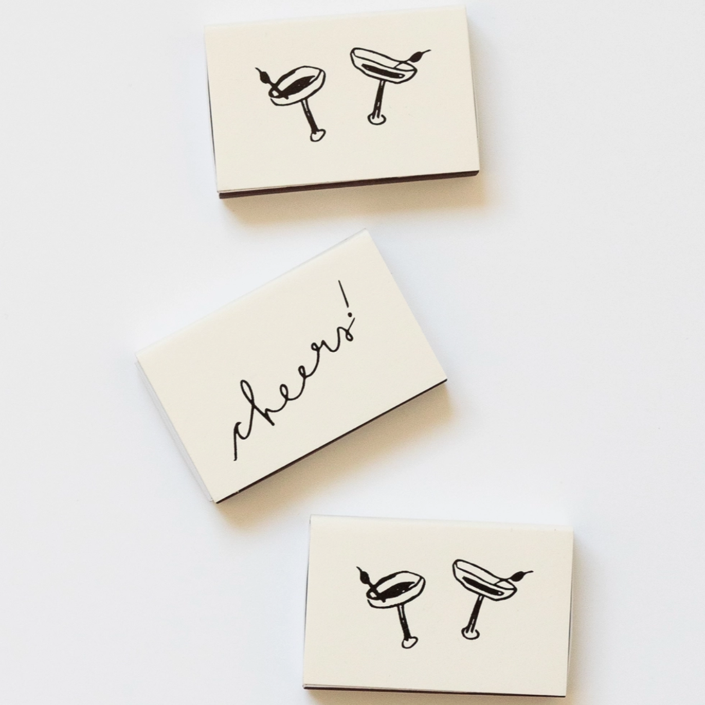 off white matchbox with black illustrations of martini glasses and the word cheers! written in script that looks like hand writing. 