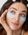 close up of womans face wearing light blue eye masks under her eyes. the masks say SOMETHING BLUE in white letters. 