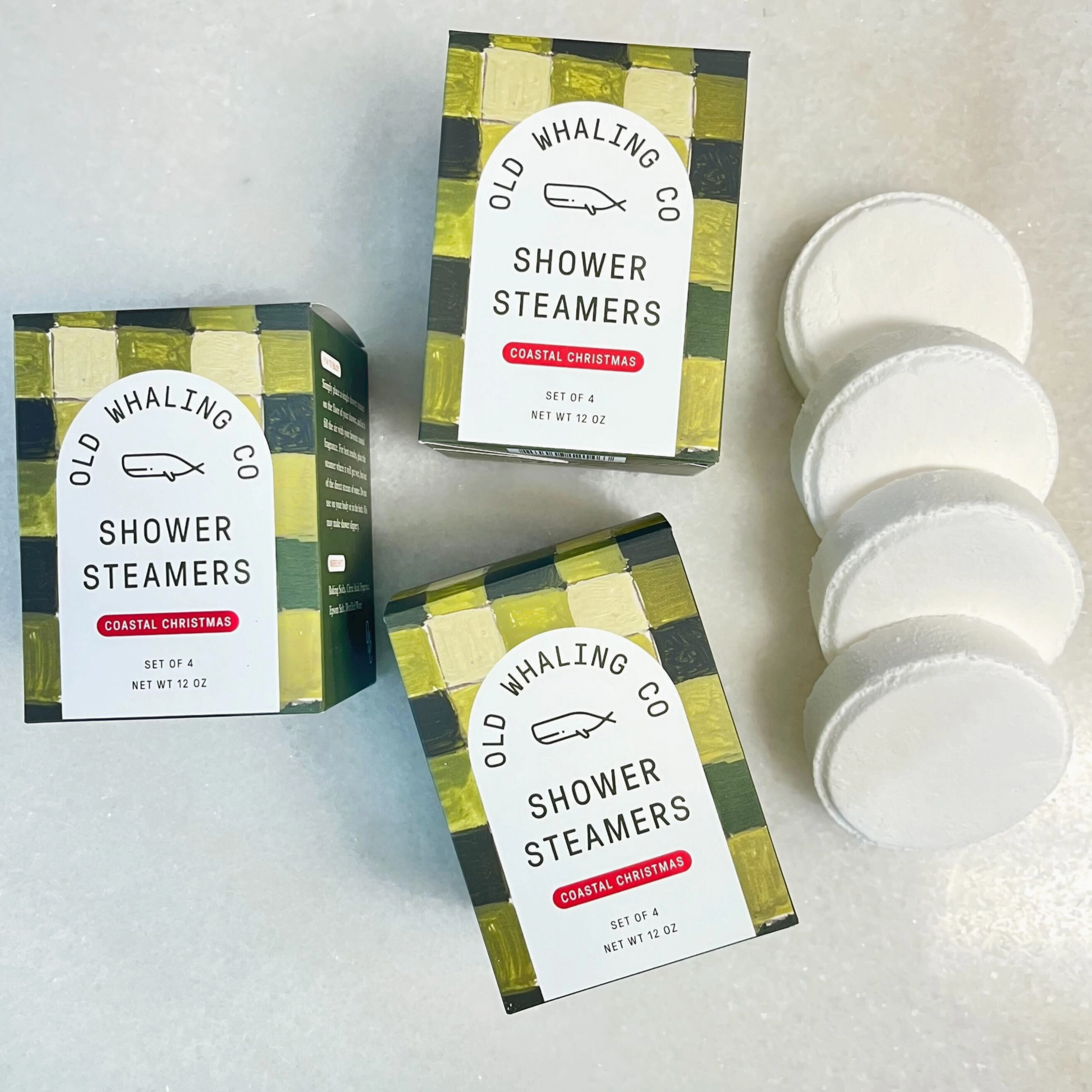 Three boxes of "Old Whaling Co Shower Steamers" in the "Coastal Christmas" scent, arranged on a light surface. Next to the boxes is a stack of four white, round shower steamers. The packaging showcases a whale logo and a mix of green, black, and gold patterns.