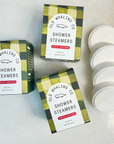 Three boxes of "Old Whaling Co Shower Steamers" in the "Coastal Christmas" scent, arranged on a light surface. Next to the boxes is a stack of four white, round shower steamers. The packaging showcases a whale logo and a mix of green, black, and gold patterns.