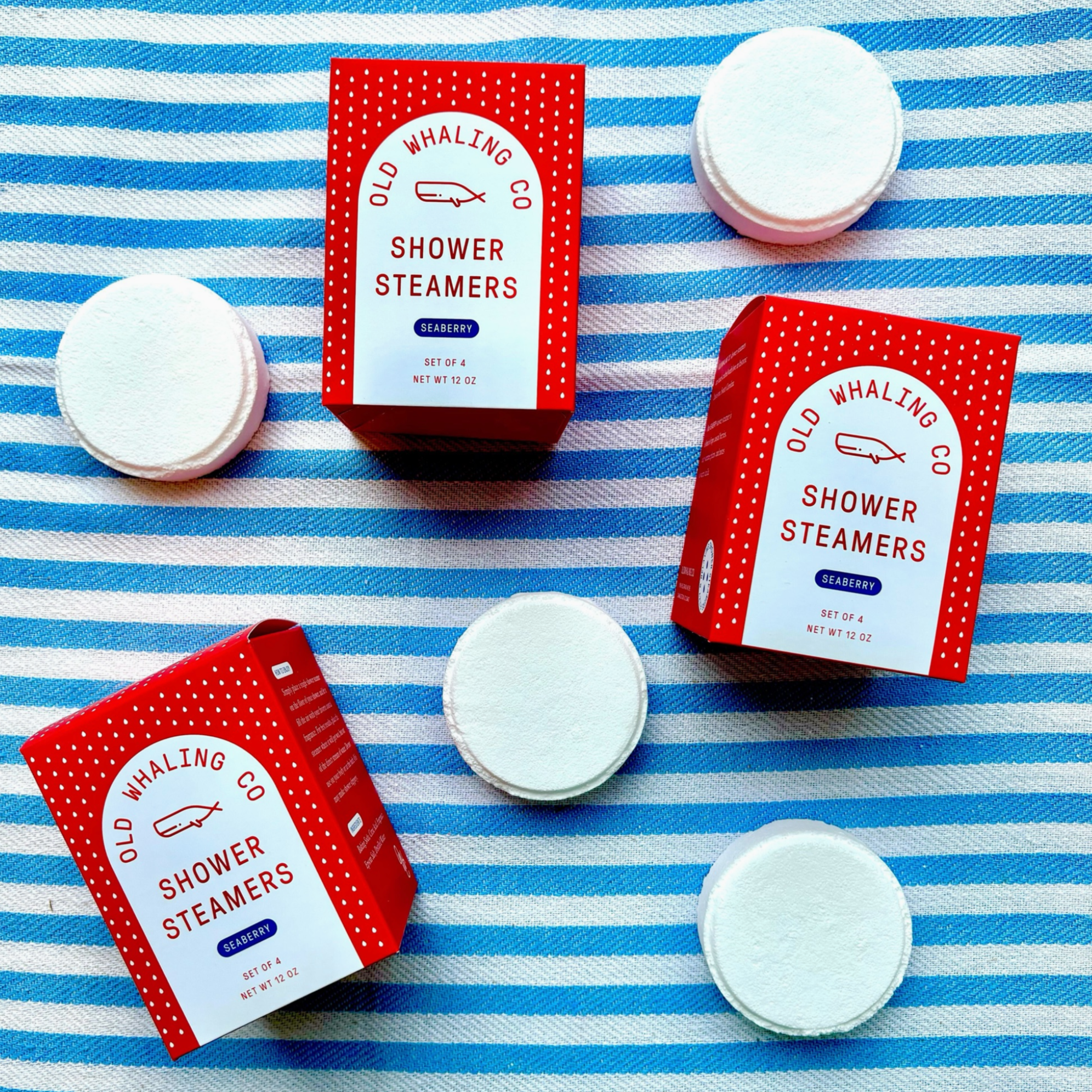 The image features several red boxes labeled "Old Whaling Co. Shower Steamers" with the scent "Seaberry." The boxes are arranged on a blue and white striped surface, accompanied by individual white, round shower steamers scattered around. The packaging displays a dotted pattern along the edges, and the front of each box mentions that it contains a "Set of 4" steamers with a net weight of 12 oz. Some parts of other products, like a red bath bomb box, are partially visible at the edges of the frame. 