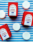 The image features several red boxes labeled "Old Whaling Co. Shower Steamers" with the scent "Seaberry." The boxes are arranged on a blue and white striped surface, accompanied by individual white, round shower steamers scattered around. The packaging displays a dotted pattern along the edges, and the front of each box mentions that it contains a "Set of 4" steamers with a net weight of 12 oz. Some parts of other products, like a red bath bomb box, are partially visible at the edges of the frame. 