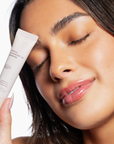 The image shows a person with radiant, glowing skin holding a tube of "ESW Beauty Vanilla Almond Butter Smoothie" lip treatment next to their face. The individual has a soft smile with glossy lips, emphasizing the product's moisturizing effect. Their eyes are gently closed, conveying a sense of calm and satisfaction. The tube, in a soft beige color, highlights ingredients like shea butter and vitamin E, designed to nourish and protect the lips. The overall mood of the image is serene and luxurious