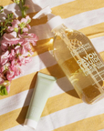 A bottle of "Compagnie de Provence Liquid Marseille Soap" placed on a yellow and white striped cloth. There are pink flowers and green stems arranged beside the bottle. The liquid soap has a clear, golden hue, and a smaller tube of hand cream from the same brand is also visible