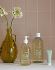 Three Compagnie de Provence products arranged on a countertop against a backdrop of light pink tiles. On the left, a brown glass vase holds two white flowers with green stems. Next to the vase are two bottles of Liquid Marseille Soap—one with a pump dispenser and a larger refill bottle. A small light green tube of Compagnie de Provence hand cream stands on the right. The arrangement gives a minimal, elegant look with soft, natural tones