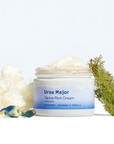 The image shows a skincare product labeled "Ursa Major Alpine Rich Cream." The jar is partially open, revealing a thick, creamy texture inside. The product highlights key ingredients on its label: cloudberry, ceramides, and tremella. Surrounding the jar are natural elements, including a white, cloud-like substance (possibly representing tremella, a type of mushroom) and green moss, reinforcing the product's natural and alpine-inspired theme.



