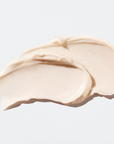 The image shows a smooth, creamy texture of a skincare product, likely a moisturizer or cream. The product has a soft, off-white color and is spread in two swipes on a light background, showcasing its rich and thick consistency.






