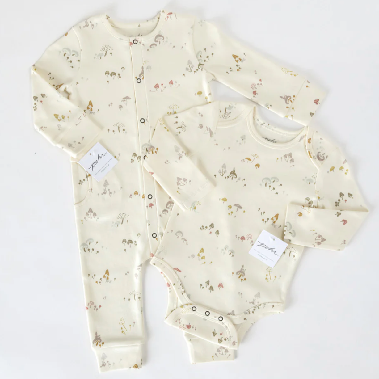 A matching baby clothing set consisting of a cream-colored long-sleeve onesie and a footed one-piece sleeper, both with whimsical mushroom and forest-themed prints. The outfits are laid flat on a white background, with small brand tags attached.