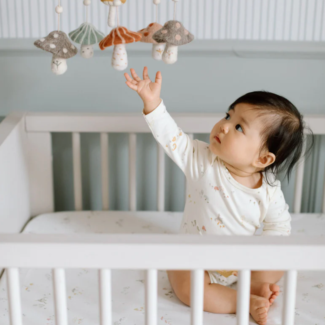 A baby wearing a cream-colored long-sleeve onesie with a mushroom and forest-themed print sits in a white crib. The baby reaches up toward a felt mobile with hanging mushroom shapes in muted colors. The room has a calm, neutral aesthetic.