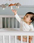 A baby wearing a cream-colored long-sleeve onesie with a mushroom and forest-themed print sits in a white crib. The baby reaches up toward a felt mobile with hanging mushroom shapes in muted colors. The room has a calm, neutral aesthetic.