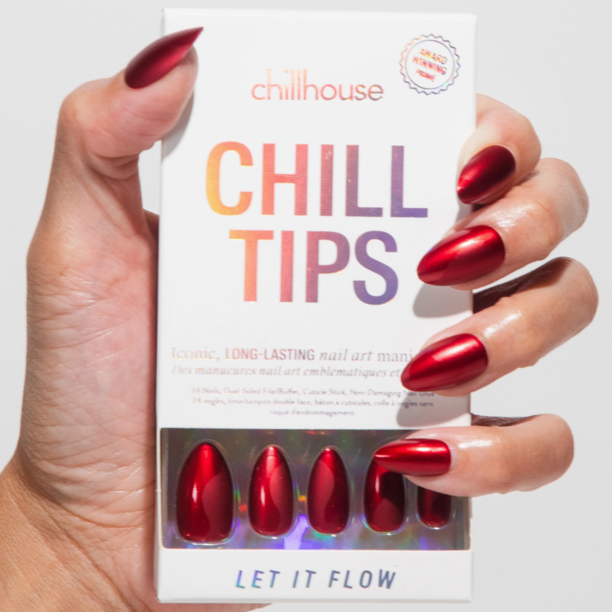 A hand with the same metallic red nails holding a box labeled "Chill Tips" from Chillhouse. The packaging shows an ombre-style logo and contains a preview of the red press-on nails.