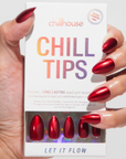 A hand with the same metallic red nails holding a box labeled "Chill Tips" from Chillhouse. The packaging shows an ombre-style logo and contains a preview of the red press-on nails.