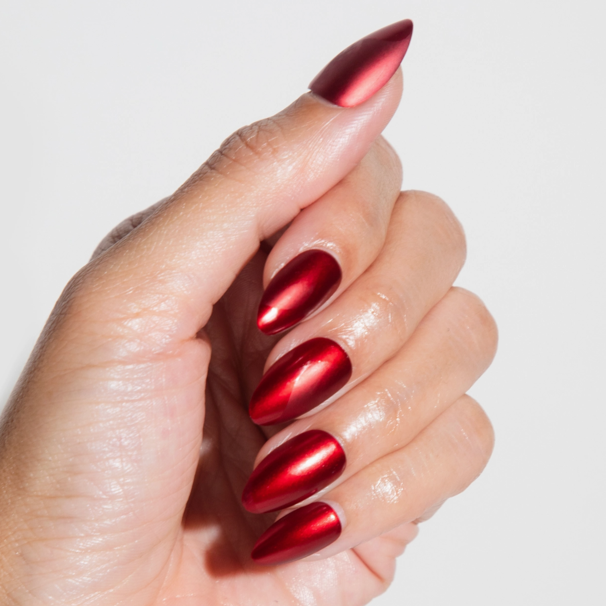 A close-up of a hand with almond-shaped nails coated in a reflective metallic red polish, giving a sleek and glossy appearance.