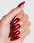 A close-up of a hand with almond-shaped nails coated in a reflective metallic red polish, giving a sleek and glossy appearance.