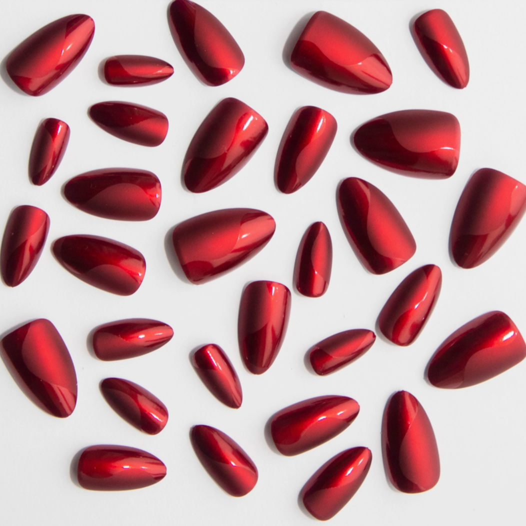 A collection of glossy, metallic red press-on nails of varying sizes scattered on a white surface. The nails are almond-shaped with a reflective finish, creating a sleek and polished look.